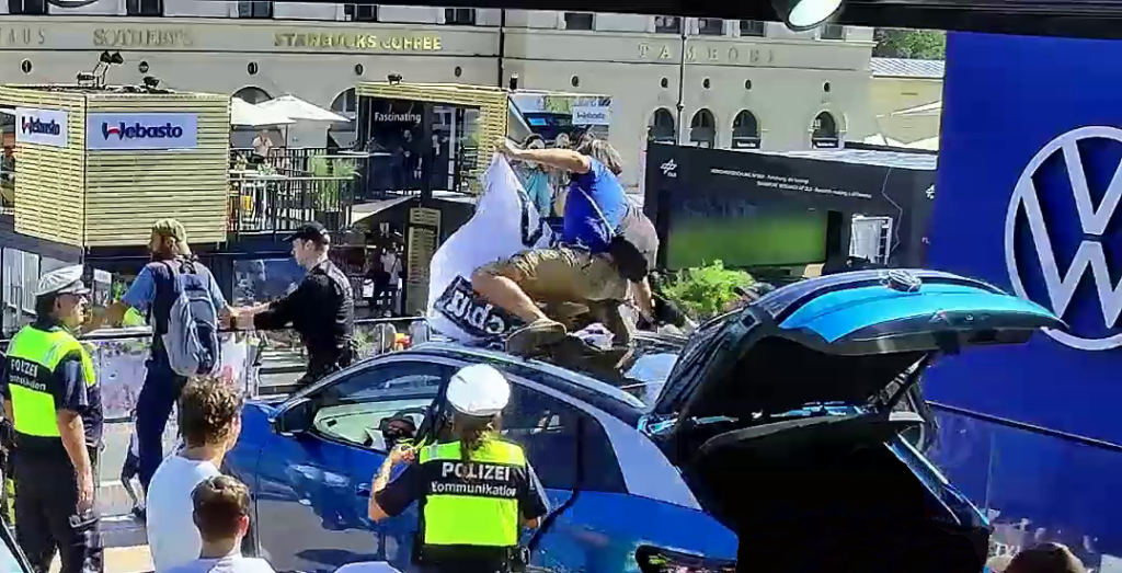Zwei Protestierende mit Bannern auf einem Volkswagen-Stand die von Polizist:innen entfernt werden